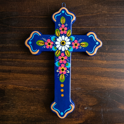 A hand-painted ceramic cross with a vibrant blue background and colorful floral designs, displayed on a dark wood surface.