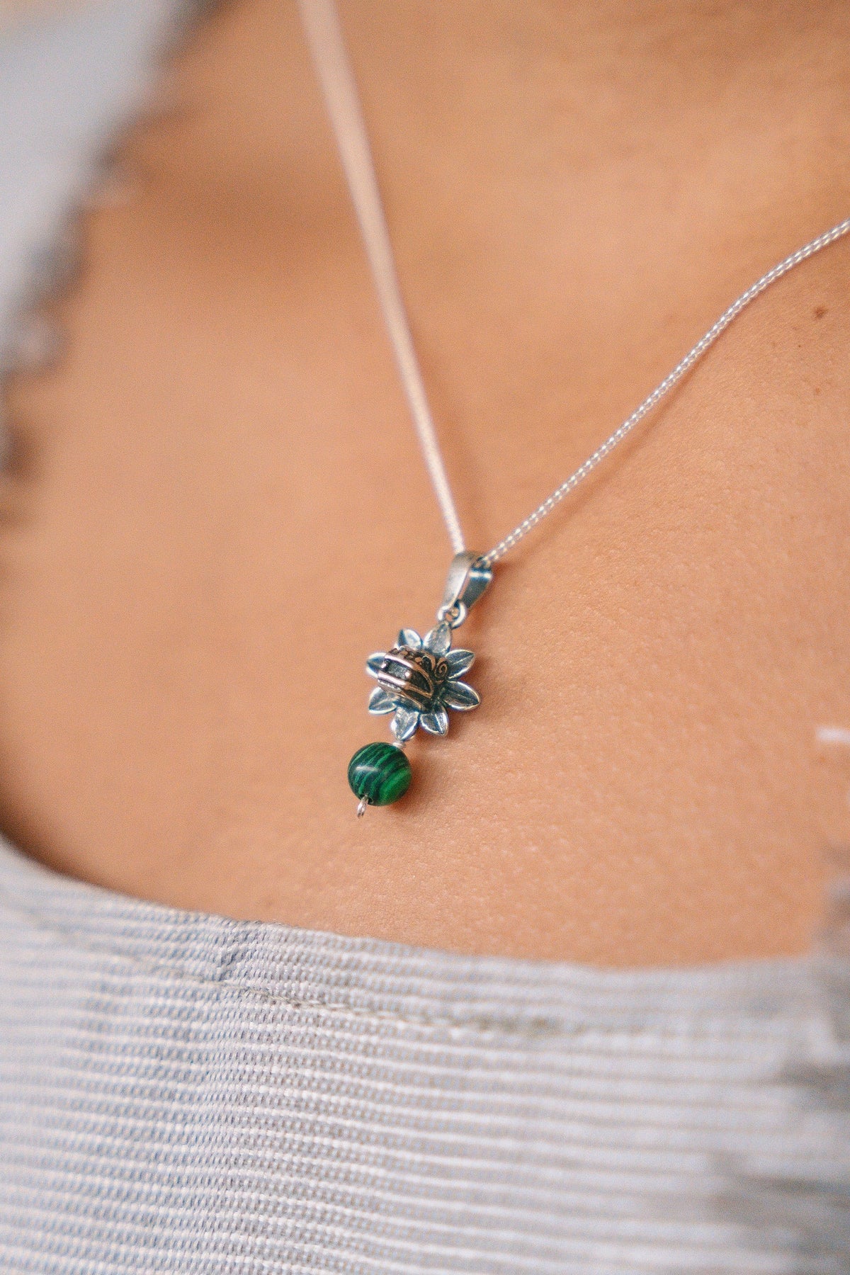 A close-up of a delicate silver chain necklace worn on a person's neck, featuring a silver flower-shaped pendant with a small green bead dangling below, set against a light gray ribbed top.