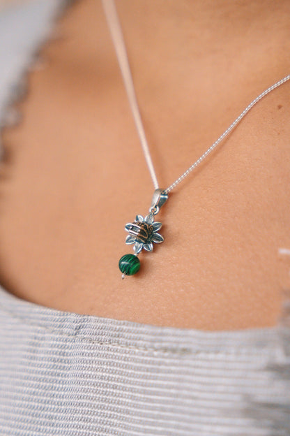 A close-up of a delicate silver chain necklace worn on a person's neck, featuring a silver flower-shaped pendant with a small green bead dangling below, set against a light gray ribbed top.