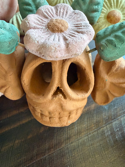 A close-up of a decorative clay skull centerpiece with a pale pink flower on top, flanked by teal and yellow flowers, set on a dark wooden surface.