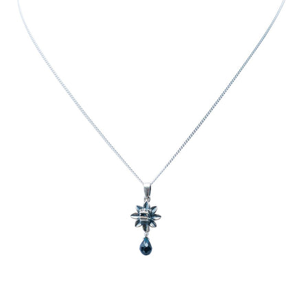 A silver-toned chain necklace featuring a starburst pendant with a dangling teardrop-shaped dark blue gemstone, set against a plain white background.