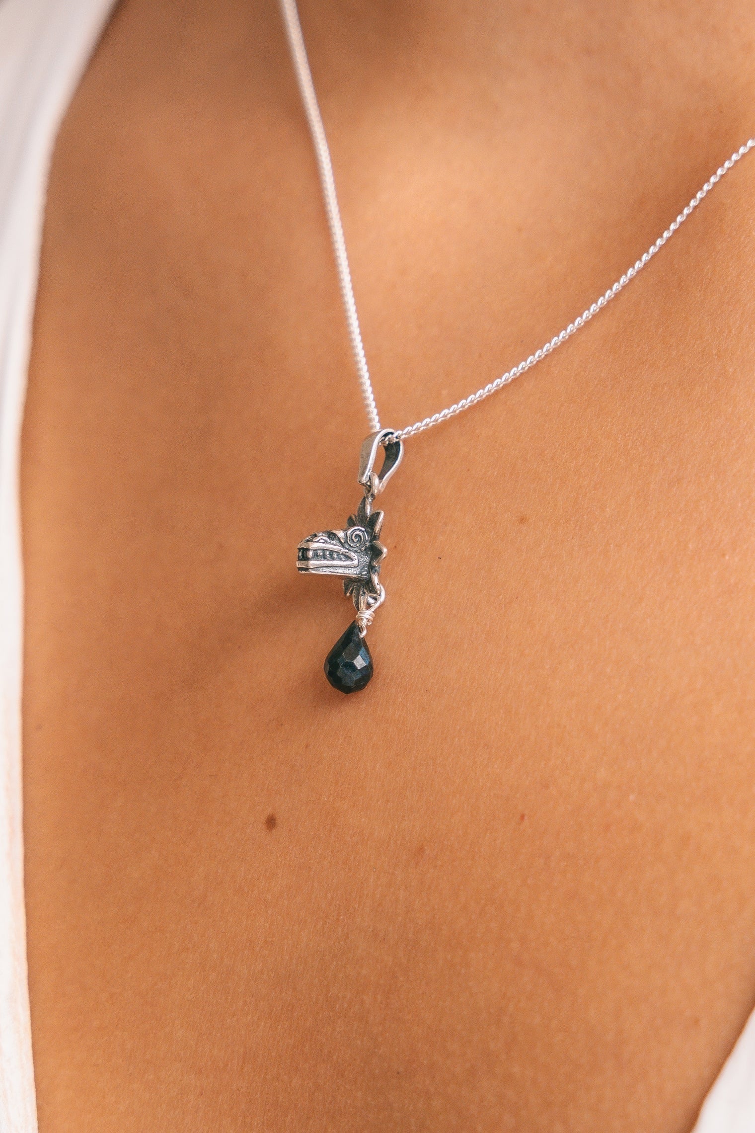 A close-up of a silver cross pendant necklace with a small dark green teardrop charm, worn on a model's neck.
