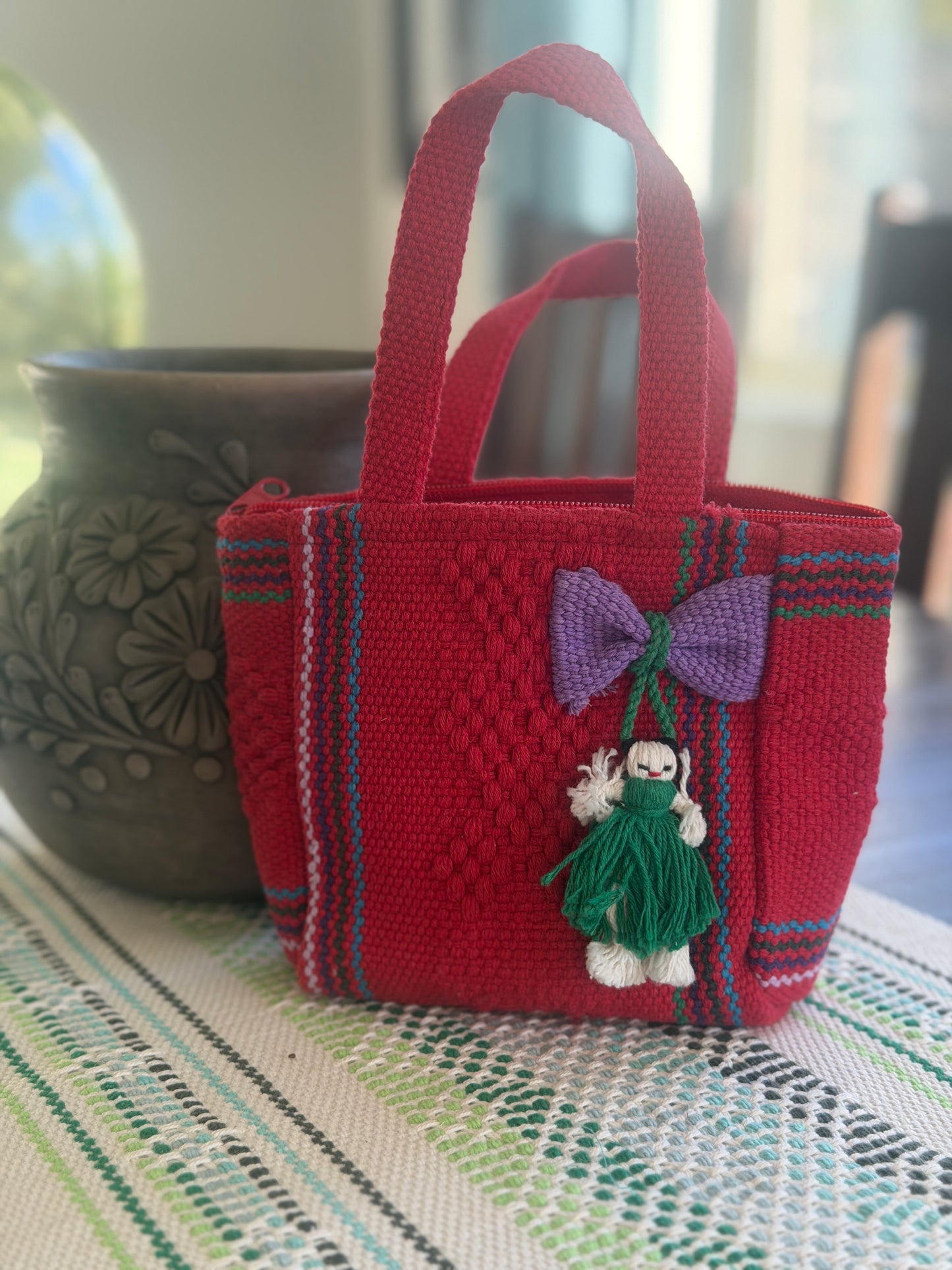 A small woven red tote bag with purple and green striped accents, featuring a purple bow and a green-and-white yarn doll charm, displayed on a patterned table runner with a blurred ceramic pot in the background.