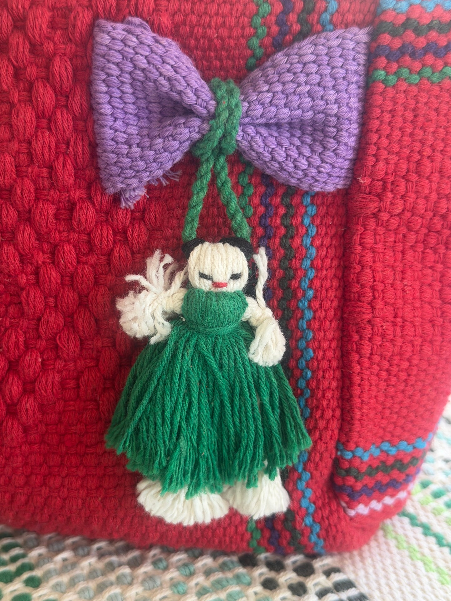 A close-up of a red knitted textile bag or basket featuring a purple knitted bow and a hanging green-and-white yarn doll ornament.