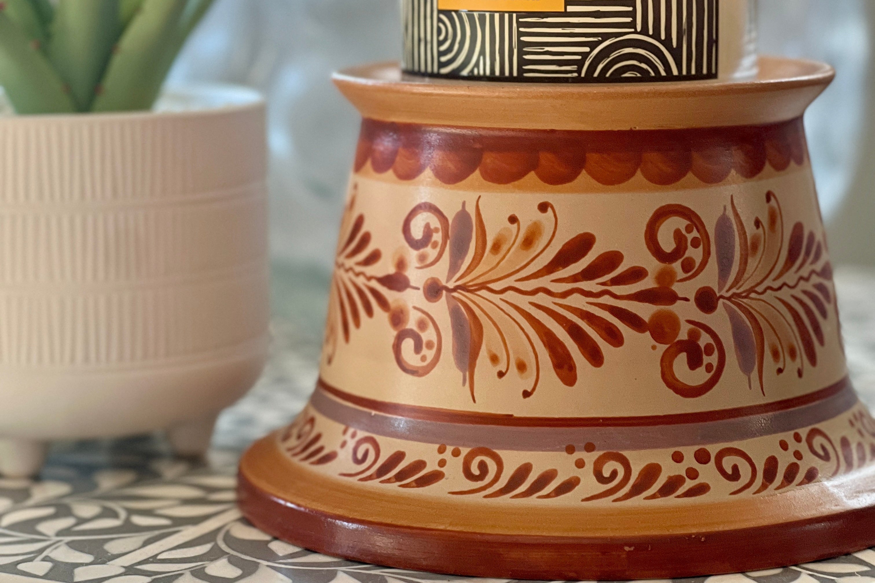 Candle with decorative label on a patterned candle holder, with a plant in the background.