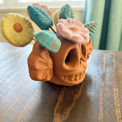 A terracotta skull-shaped planter with a floral crown of pastel flowers and leaves, sitting on a dark wooden tabletop.