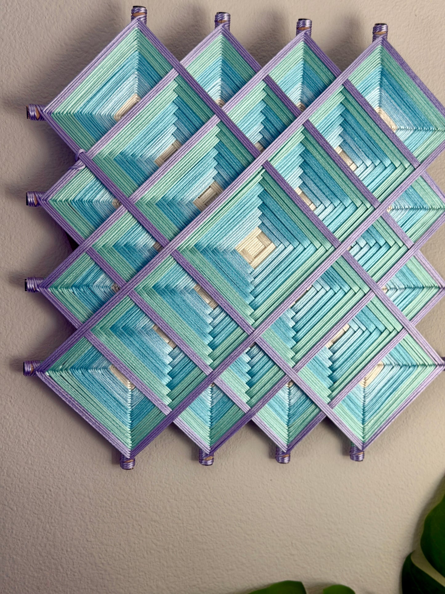 A geometric wall hanging made of layered diamond panels in shades of blue, teal, and lavender, mounted on a beige wall.