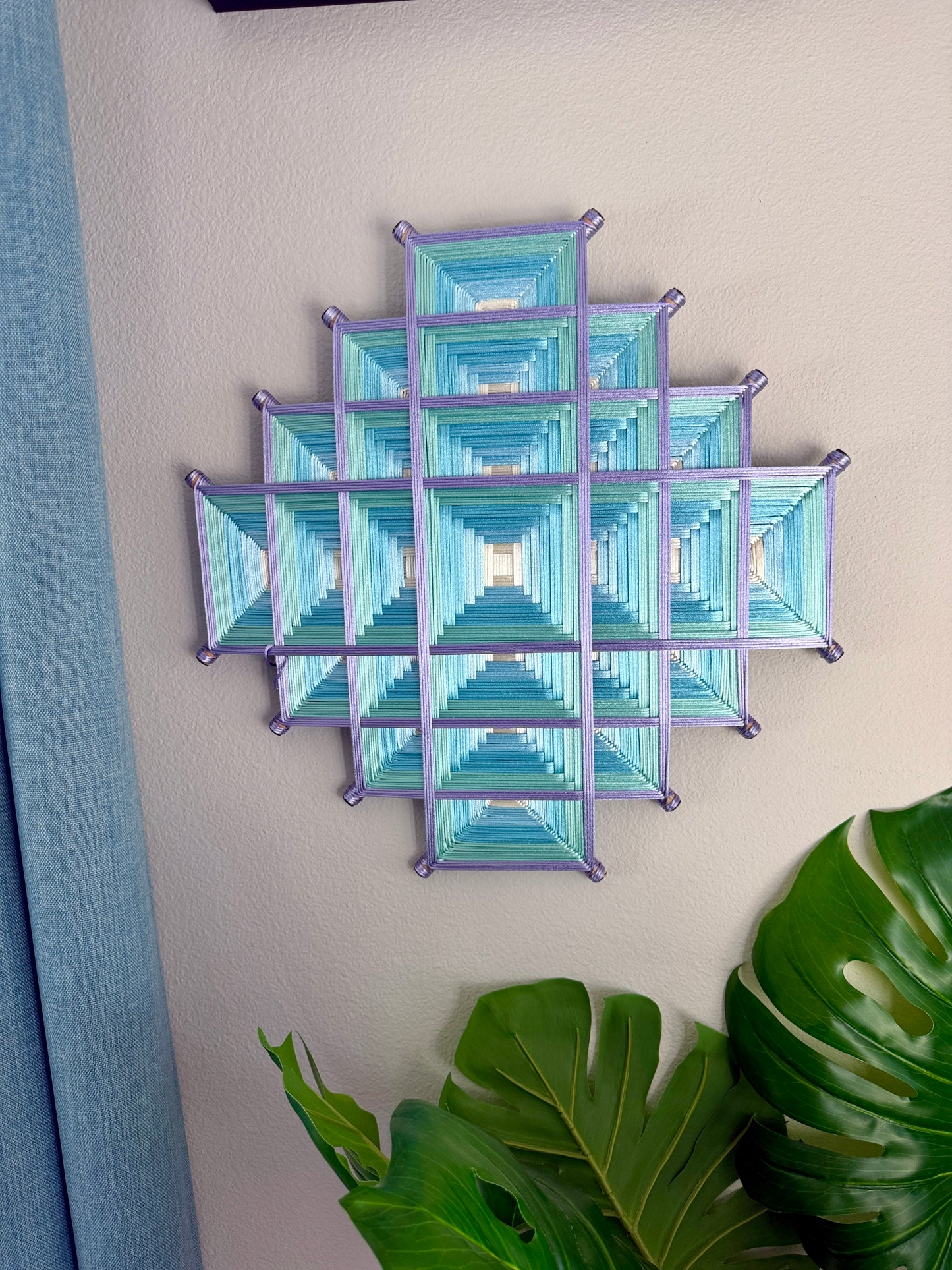 A geometric wall art piece made of layered blue and green striped triangles arranged in a diamond pattern, mounted on a beige wall with a blue curtain and green plant leaves visible.