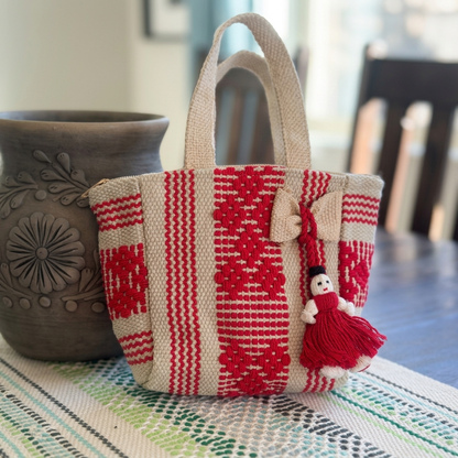 A woven tote bag with red and beige geometric patterns, featuring two top handles and a knitted red dress doll charm, set on a patterned table runner next to a dark ceramic vase.