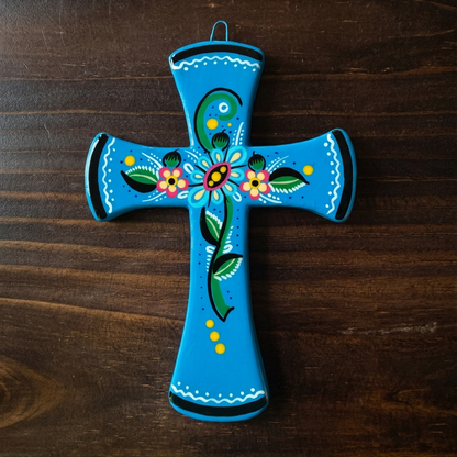 A hand-painted ceramic cross in bright turquoise blue with colorful floral and paisley designs, resting on a dark brown wooden surface.