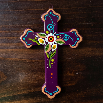 A hand-painted Ceramic cross with a deep purple background, decorated with a central white flower, green leaves, and turquoise and orange accents, resting on a dark wood surface.