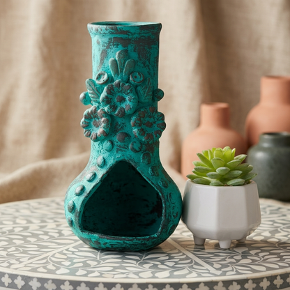 A distressed teal ceramic vase with floral relief details and a hollow base, displayed on a patterned table next to a small potted succulent with blurred terracotta vases in the background.