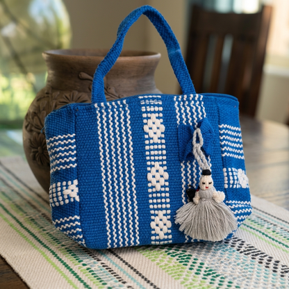 A blue and white woven tote bag with a gray tassel charm featuring a small doll, displayed on a patterned runner in front of a rustic brown ceramic pot.