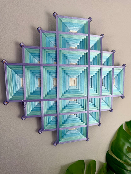 A geometric wall hanging made of multiple square panels with layered teal, blue, and mint striped triangles, outlined in purple, mounted on a beige wall with green leaves visible at the bottom right.