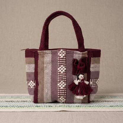 A small woven tote bag with dark burgundy handles and trim, featuring vertical striped panels in burgundy, beige, and gray, and a hanging doll charm, set against a neutral textured background.