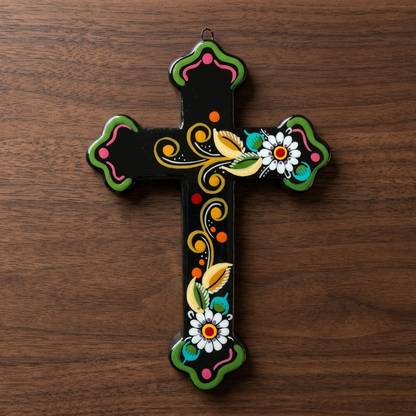 A hand-painted ceramic cross with a black base, decorated with green, yellow, pink, white, and orange folk art motifs, resting on a dark brown wooden surface.