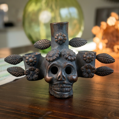 A dark metallic gothic skull candle holder with floral and leaf details, displayed on a wooden table with a blurred green glass vase in the background.