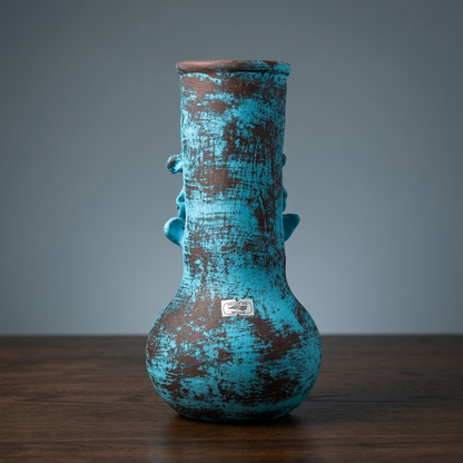 A rustic ceramic vase with a distressed turquoise and brown glaze, featuring small side handles and a small silver brand plaque, set on a dark wooden surface against a gray background.