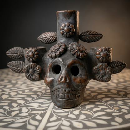 A dark brown ceramic Day of the Dead style skull candle holder with floral and leaf details, displayed on a patterned table against a dark background.