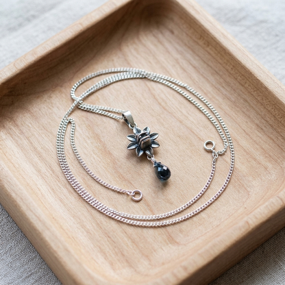 A silver-toned necklace with a flower-shaped pendant and a small dark teardrop bead, displayed on a light wood tray over a neutral fabric background.