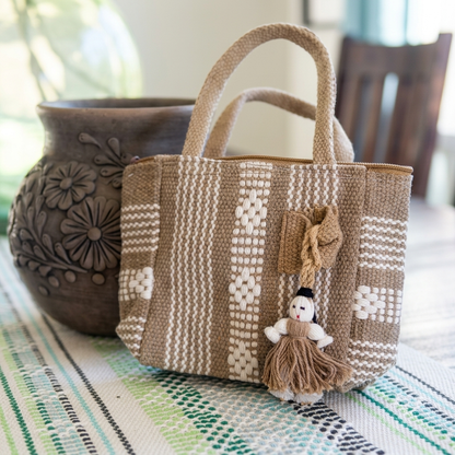A woven tote bag in natural beige and cream with geometric patterns, featuring dual top handles and a small doll charm, displayed on a patterned table runner next to a dark ceramic pot.