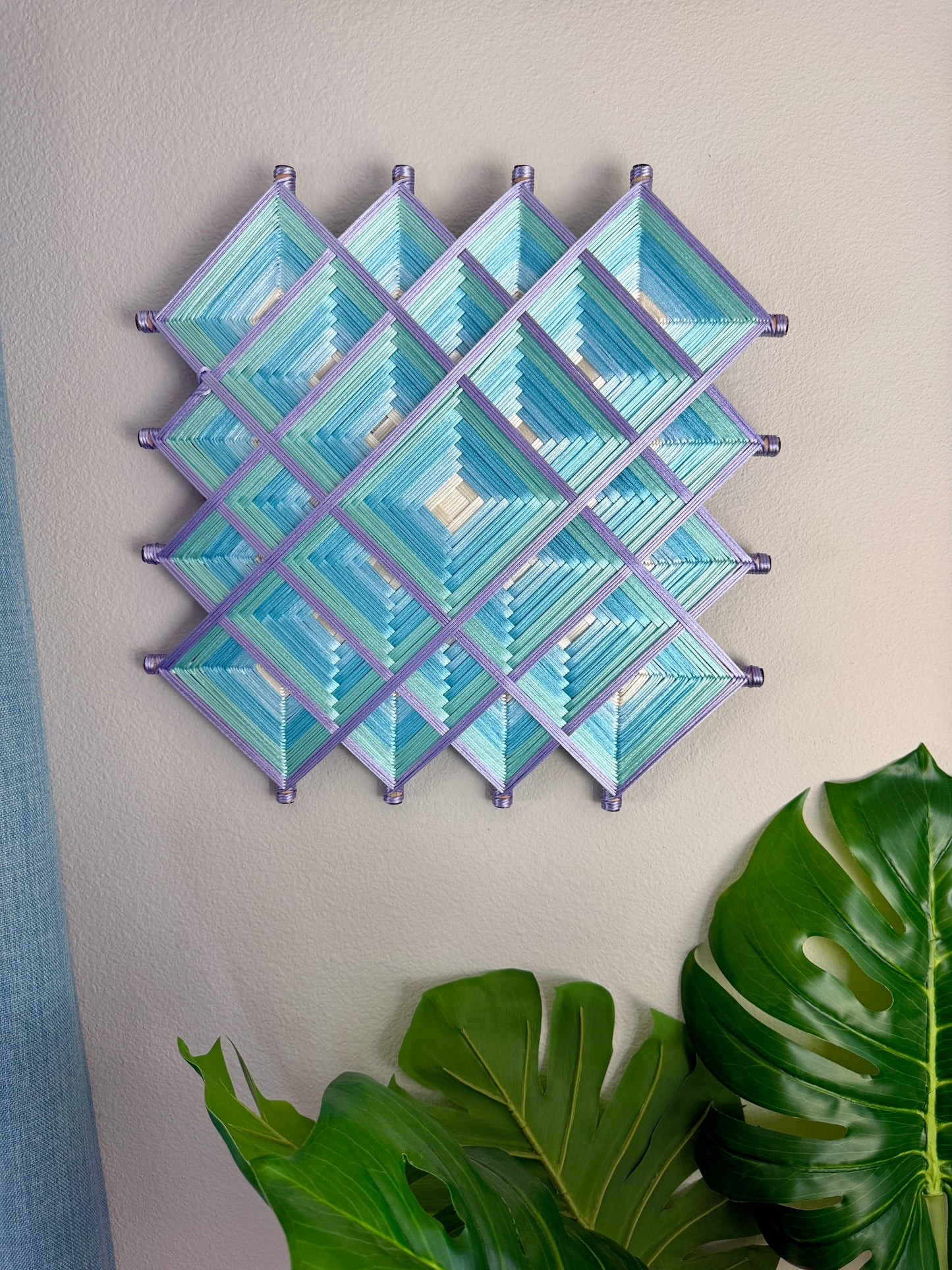 A geometric wall hanging made of layered diamond and triangle shapes in shades of light blue and teal, outlined with lavender-purple borders, displayed on a beige wall with green plant leaves in the foreground.