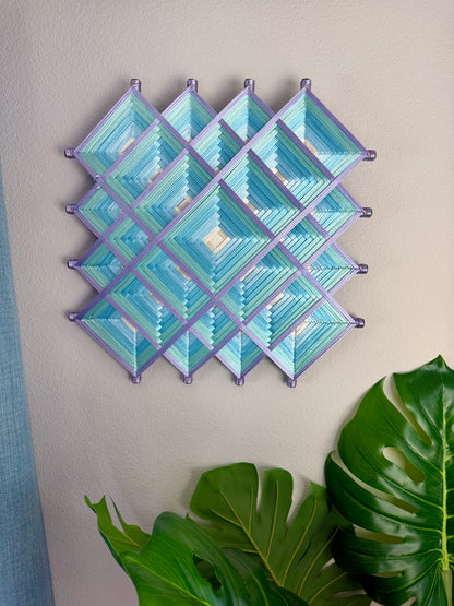 A geometric wall hanging made of layered diamond and triangle shapes in shades of light blue and teal, outlined with lavender-purple borders, displayed on a beige wall with green plant leaves in the foreground.