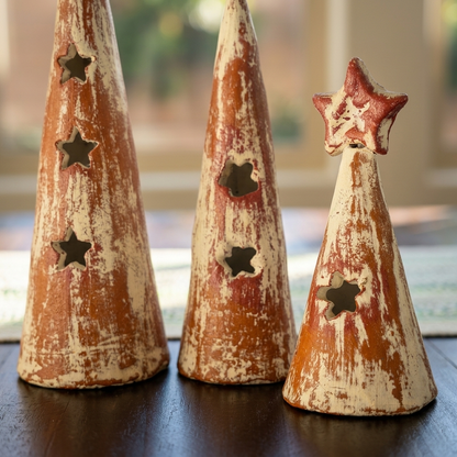 Three distressed ceramic Christmas tree-shaped candle holders with star cutouts, displayed on a dark wooden surface in a softly lit indoor setting.