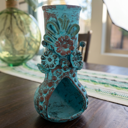 A turquoise and brown distressed ceramic vase with floral relief details, displayed on a dark wooden table in a home setting.