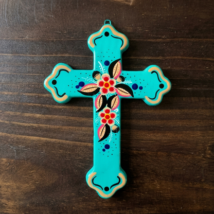 A turquoise hand-painted wooden cross with floral and geometric folk art designs, displayed on a dark wood background.
