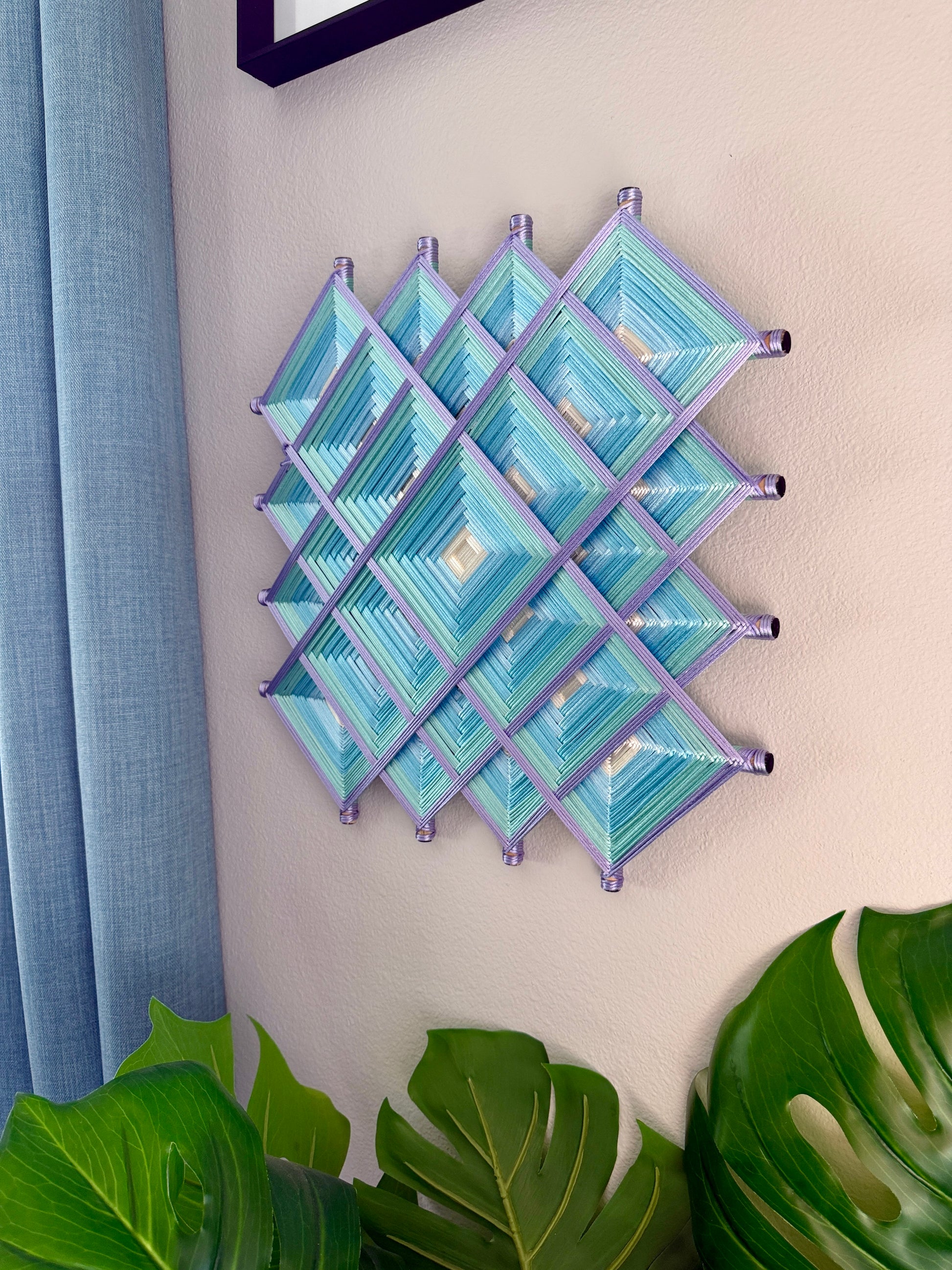 A decorative wall hanging made of interlocking diamond-shaped panels with layered blue, teal, and light purple striped patterns, mounted on a beige wall with a blue curtain and green leaves visible at the sides.
