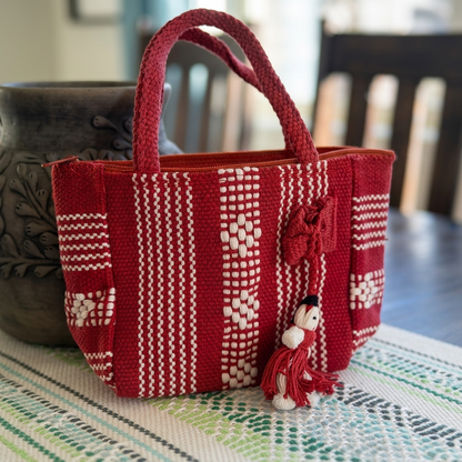 A red and white handwoven tote bag with braided handles and a small decorative figure charm, placed on a patterned table runner in a home setting.