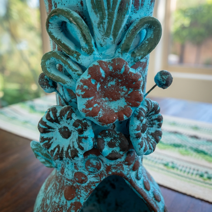 A close-up of a decorative ceramic vase with a distressed turquoise and brown patina finish, featuring floral and scroll motifs, set on a wooden table with a blurred indoor background.