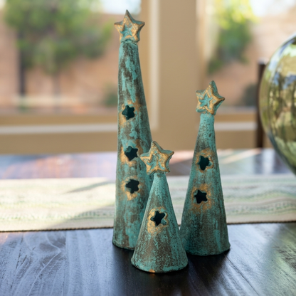 Three decorative metal Christmas tree ornaments with a distressed turquoise and gold finish, displayed on a dark wooden table in a home setting.