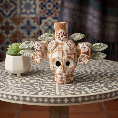 A decorative ceramic skull vase with floral details and leaf-like side sprigs, displayed on a patterned round table next to a small potted succulent.