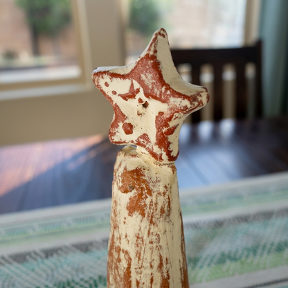 A rustic, hand-painted wooden star-shaped ornament with a weathered cream and reddish-brown finish, standing on a patterned table runner in a home setting.