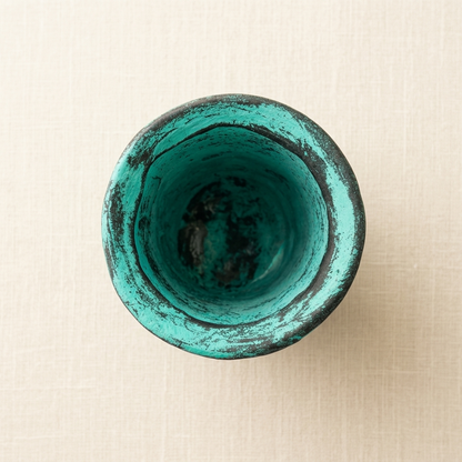 A top-down view of a small ceramic cup or bowl with a distressed turquoise green glaze and a dark rim, set against a light beige textured background.