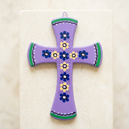 A hand-painted purple ceramic cross ornament with blue, yellow, and green floral patterns, displayed on a light beige stone block.